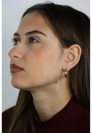 Jeune femme aux longs cheveux bruns, portant un col roulé bordeaux et une boucle d'oreille en or en forme d'anneau, regardant vers le haut sur un fond uni.