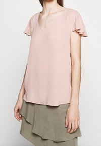 Jasnoróżowa bluzka z krótkimi falbanami, dekoltem w kształcie V, miękką fakturą i luźnym krojem, zestawiona z warstwową oliwkową spódnicą.