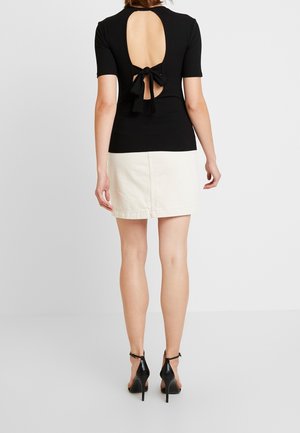 Kobieta ubrana w czarną bluzkę z krótkim rękawem i odkrytymi plecami, zawiązaną kokardą, białą mini spódniczkę oraz czarne szpilki z paskiem na kostce, stojąca.