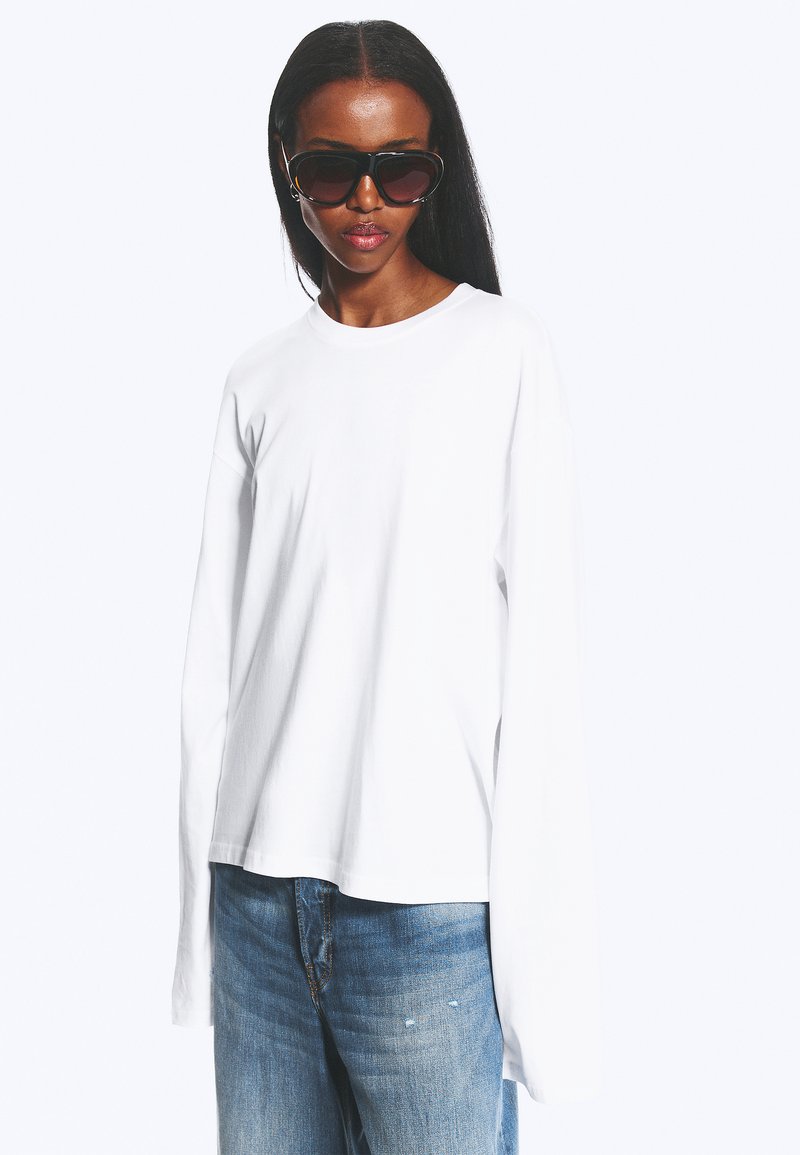 Mujer con cabello largo que lleva gafas de sol negras de gran tamaño, camisa blanca de manga larga y vaqueros azules sobre un fondo liso.