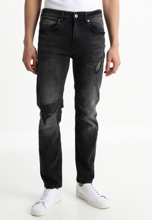 Person trägt ausgewaschene schwarze zerrissene Jeans mit gerippten Aufnähereien und weiße Turnschuhe und steht vor einem einfarbigen weißen Hintergrund.