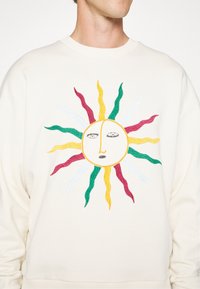 Cremefarbener Sweatshirt mit einem bestickten Sonnenmotiv mit Gesicht. Strahlen in Grün, Gelb, Rot und Weiß. Weiche Textur und lockere Passform.