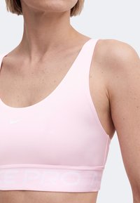 Γυναίκα που φοράει ροζ αθλητικό σουτιέν Nike με στρογγυλή λαιμόκοψη και φαρδιές τιράντες, που κόβεται στους ώμους και στο πάνω μέρος του στήθους.