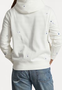 Vit sweatshirt med huva, prydd med blå färgfläckar på baksidan. Mjukt tyg, åtsittande ärmar och ribbad nederkant. Bärs med blå jeans.