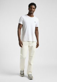 T-shirt en coton blanc avec un logo "Lee" brodé, associé à un jean couleur crème et des baskets grises, montrant une tenue décontractée.