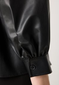 Zwarte faux leren blouse met puffmouwen, voorzien van een zachte textuur en knoopmanchetten. Close-up details benadrukken de glans van het materiaal.