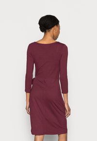 Vestido bordô com mangas três-quartos, design ajustado, detalhe de laço na cintura e textura suave, atingindo logo acima do joelho.