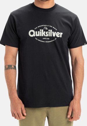 Mand iført sort Quiksilver T-shirt med hvidt logo i midten, kombineret med olivengrønne bukser og en tribal tatovering på venstre underarm.