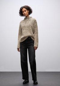 Beżowy sweter w wzory z wysokim kołnierzem, teksturowana tkanina, w zestawieniu z czarnymi spodniami wysoki stan i czarnymi botkami na kostkę, stojący na szarym podłodze.