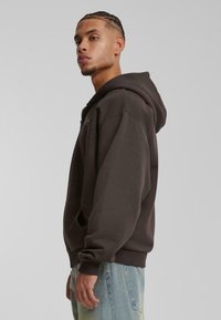 Sudadera con capucha marrón, con un bolsillo delantero, puños y dobladillo acanalados. Lleva puesta unos vaqueros de color azul claro, con un diseño minimalista.
