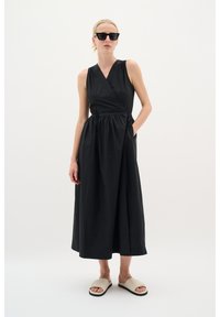 Schwarzes ärmelloses Wickelkleid mit taillierter Taille und Taschen, aus leichtem Stoff, der bis zur Mitte der Waden reicht; kombiniert mit beigen Rutsch-Sandalen.