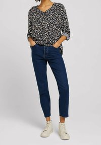 Blouse noire avec motifs abstraits beiges et blancs, col en V, manches retroussées ; portée avec un jean bleu raccourci et des baskets blanches montantes.