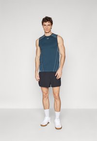 Ermeløs teal sportskjorte med grå detaljer og hvit søm, kombinert med svarte shorts. Modellen har på seg hvite sneakers og ankelsokker.