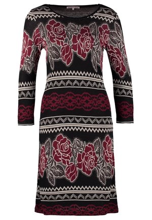 Robe noire à motif floral avec des manches trois-quarts, associée à un sac fourre-tout en cuir noir. Le mannequin porte des escarpins noirs.