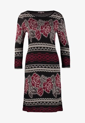 Vestido negro con estampado floral y mangas tres cuartos, combinado con un bolso tote de cuero negro. La modelo lleva zapatos de tacón negros.