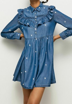 Robe en jean bleu avec broderie florale blanche, empiècement à volants, manches longues, devant boutonné, portée par une personne les mains sur les hanches.