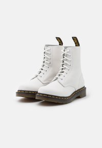 Botine de piele albă până la gleznă cu talpă neagră, cusături galbene și fâșii negre pentru prindere. Prezintă un design cu șiret și o textură de suprafață netedă.