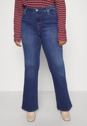 Person iført mørkeblå high-waisted bootcut jeans, stribet vinrød langærmet skjorte og beige ankelstøvler, stående mod en hvid baggrund.