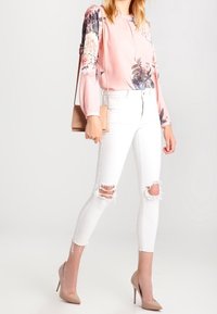 Blusa rosa a maniche lunghe con stampa floreale sulle maniche, abbinata a jeans skinny bianchi strappati e scarpe con tacco color nude. Piccola borsa a tracolla.