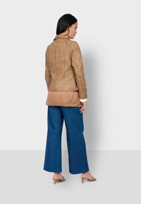 Tanlederjacke mit taillierter Passform und diagonalen Nähten, kombiniert mit weit geschnittenen blauen Jeans und nudefarbenen Absätzen, von hinten betrachtet.