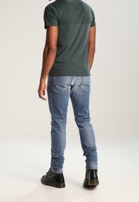 Tmavě zelené tričko s krátkým rukávem a světle zeleným panelem, doplněné světle modrými džínami a černými šněrovacími botami, pohled zezadu.