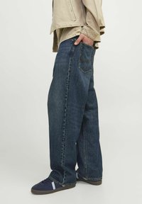 Jeans de mezclilla azul de pierna ancha con un acabado desgastado, que presentan un estilo clásico de cinco bolsillos y costuras oscuras. Combinados con zapatillas de deporte azul marino.