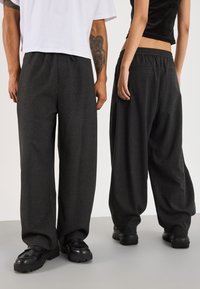 To personer i mørkegrå brede bukser og sorte sko, den ene iført en hvid skjorte, den anden en sort crop top, står på et gråt gulv.
