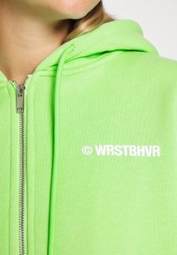 Fleece verde brilhante com fecho, feito de algodão macio, apresenta um fecho de correr prateado e o logotipo impresso em branco "WRSTBHVR" na frente. Acabamento de tecido texturizado.