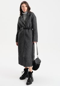 Cappotto grigio a zigzag con vita cinturata, abbinato a un dolcevita grigio. Stivali neri con lacci e dettagli in pelliccia e una piccola borsa nera.