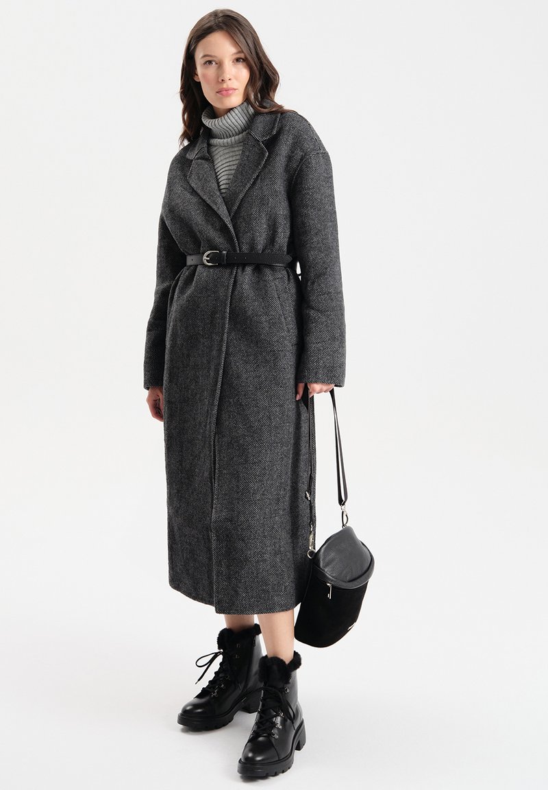 Cappotto grigio a zigzag con vita cinturata, abbinato a un dolcevita grigio. Stivali neri con lacci e dettagli in pelliccia e una piccola borsa nera.