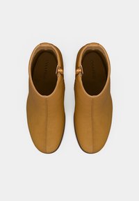 Világosbarna bőr bokacsizma sima textúrával, oldalsó cipzáras záródással és díszítő fémes kiemelésekkel. A cipő elején varrott varrás részlet.