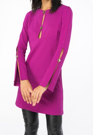 Frau trägt ein langärmeliges, magentafarbenes Kleid mit goldenen Knopfakzenten und Ärmelschlitzen, kombiniert mit glänzenden schwarzen Leggings.