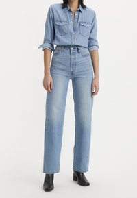 Camisa de mezclilla azul claro con dos bolsillos en el pecho, cuello abotonado, combinada con jeans de pierna ancha y tiro alto en azul claro y botines negros.