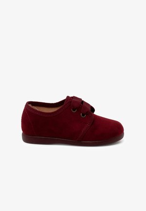 Chaussure en daim rouge avec un bout arrondi, fermeture par sangle élastique et semelle plate. Texture lisse avec un design simple et des accents de couture.