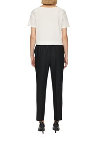 Kvinna iklädd en vit kortärmad topp, svarta högmidjade byxor och svarta klackskor, står vänd bort från kameran.