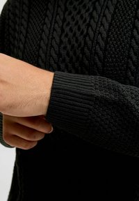 Black knit sweater featuring cable and textured patterns with a ribbed cuff; close-up of arm adjusting sleeve.