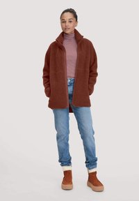 Rostfarbener Fleecejacke mit hohem Kragen, Frontreißverschluss, Seitentaschen, getragen über einem gestreiften Rollkragenpullover, kombiniert mit blauen Jeans und braunen Stiefeln.