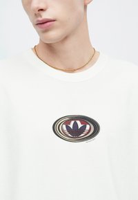 Fehér pamut póló, amelyen több színű ovális grafika látható, lila, piros és fekete árnyalatú virágos logóval. Arany lánc nyaklánc.