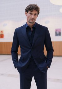 Homme aux cheveux bruns portant un costume bleu foncé et une chemise, debout à l’intérieur, les mains dans les poches et une expression neutre.