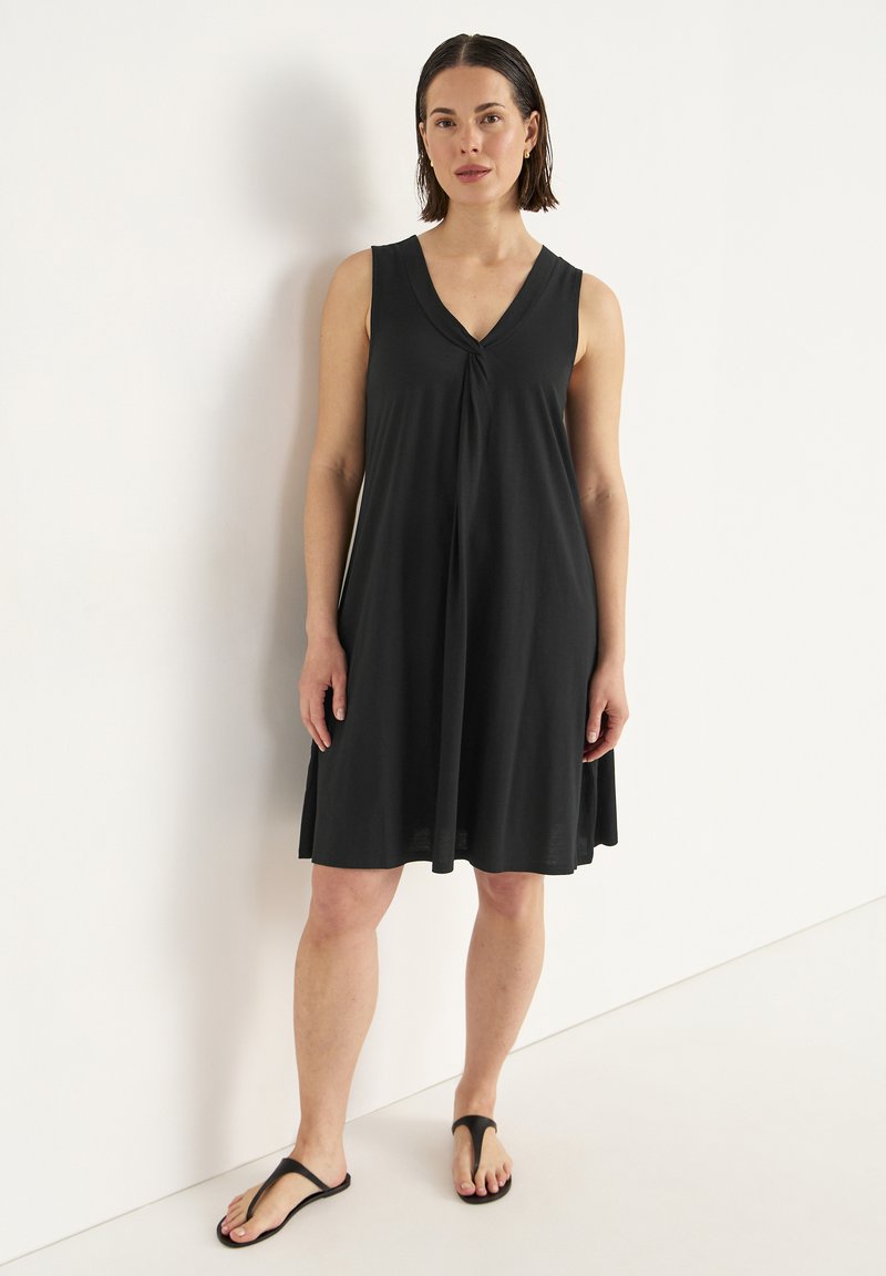 Schwarzes ärmelloses Kleid mit V-Ausschnitt und gerafften Details an der Brust. Weicher Stoff, A-Linien-Form, knielang, kombiniert mit schwarzen Flip-Flops.