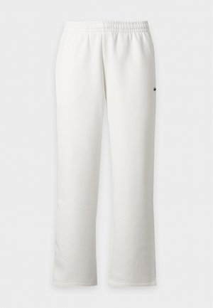 Homme debout portant une chemise noire à manches longues, un pantalon blanc et des baskets blanches sur un fond clair uni.