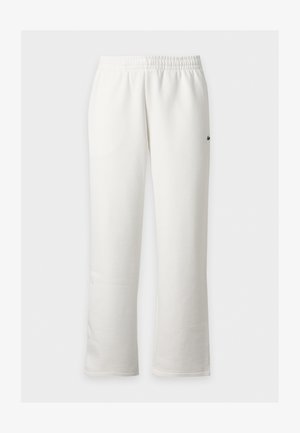 Homme debout portant une chemise noire à manches longues, un pantalon blanc et des baskets blanches sur un fond clair uni.