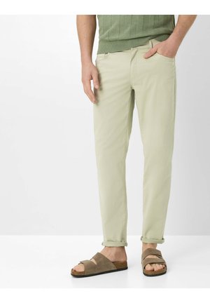 Mann trägt beigefarbene, schmal geschnittene Hose mit umgeschlagenen Säumen, grünes Strickoberteil und braune Wildledersandalen, steht vor einem schlichten hellen Hintergrund.