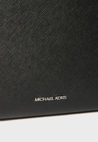 Sac en tissu noir texturé avec un logo "MICHAEL KORS" en relief argenté. Forme rectangulaire générale avec une surface légèrement texturée.