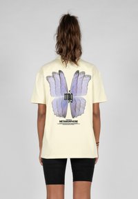Gul bomulds T-shirt med et stort sommerfuglegrafik i nuancer af lilla, med sort tekst nedenunder, der beskriver "Metamorphose."
