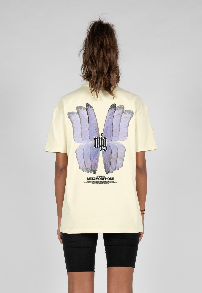 Gul bomulds T-shirt med et stort sommerfuglegrafik i nuancer af lilla, med sort tekst nedenunder, der beskriver "Metamorphose."