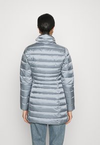 Mujer con el cabello oscuro recogido lleva una chaqueta puffer acolchada de color gris claro y jeans azules, de frente a una pared blanca lisa, de pie y erguida.