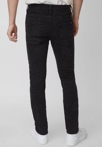Persona vistiendo jeans negros ajustados y zapatillas blancas, mostrada desde atrás contra un fondo gris claro liso.