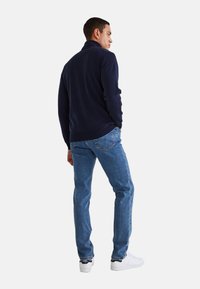 Pull en marine à col montant, porté avec un jean bleu clair et des baskets blanches. Mise au point sur la vue arrière, mettant en valeur la texture du tissu.
