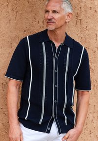 Polo de manga corta en color azul marino con rayas verticales blancas y detalles perforados, que cuenta con cuello y cierre de botones en la parte delantera.